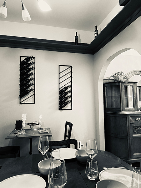 Image, Point de vue de la table assise vers l'espace ouvert. En arrière-plan, des bouteilles de vin sont accrochées au mur sur une étagère.